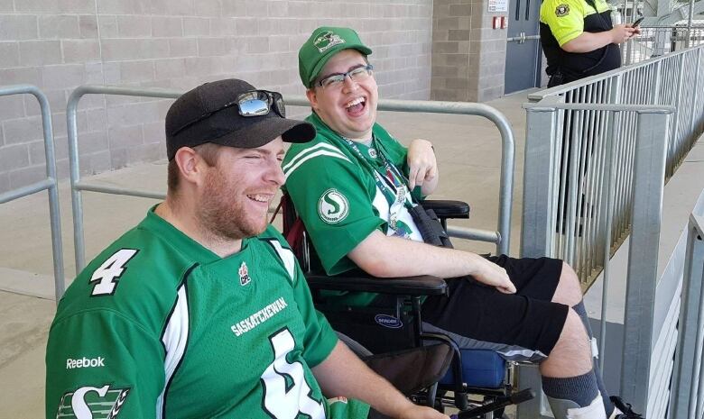 Two men in green shirts and ballcaps can be seen seated at a stadium, the one on the right in a wheelchair.