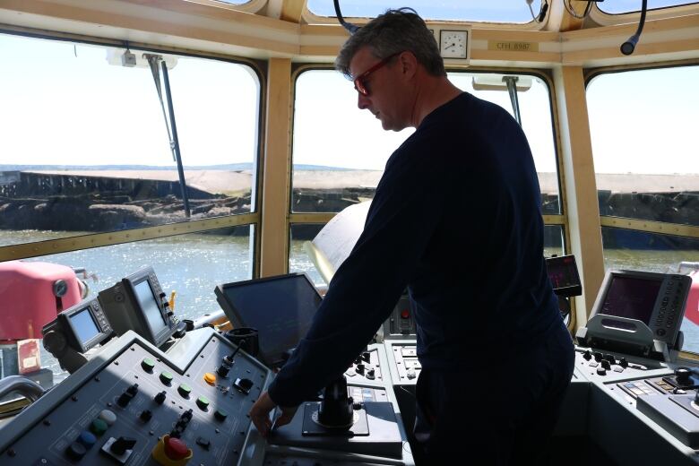 A fortyish man in sunglasses stands in the wheelhouse of a tugboat, working the controls with the harbour visible in front of him. 