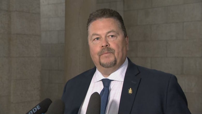 A man wearing a dark blue blazer, white shirt and blue tie looks at a reporter while answering questions in a scrum.