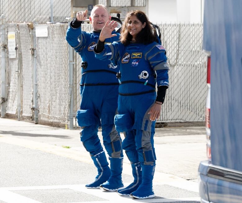 A man and woman in blue astronaut jumpsuits are seen waving.