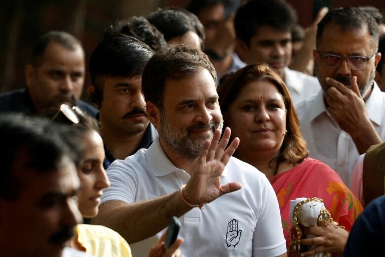 A man with a greyish beard, wearing a white shirt, waves as he walks through a crowd.