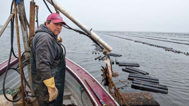 A man dressed in fishing gear stands on a boat with floating oyster boxes in the background. 