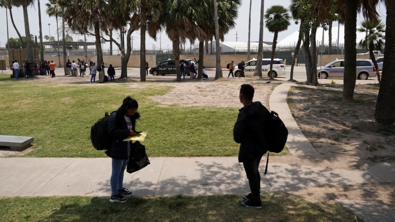 Two people carrying large backpacks stand on grass outside a government building holding paperwork.