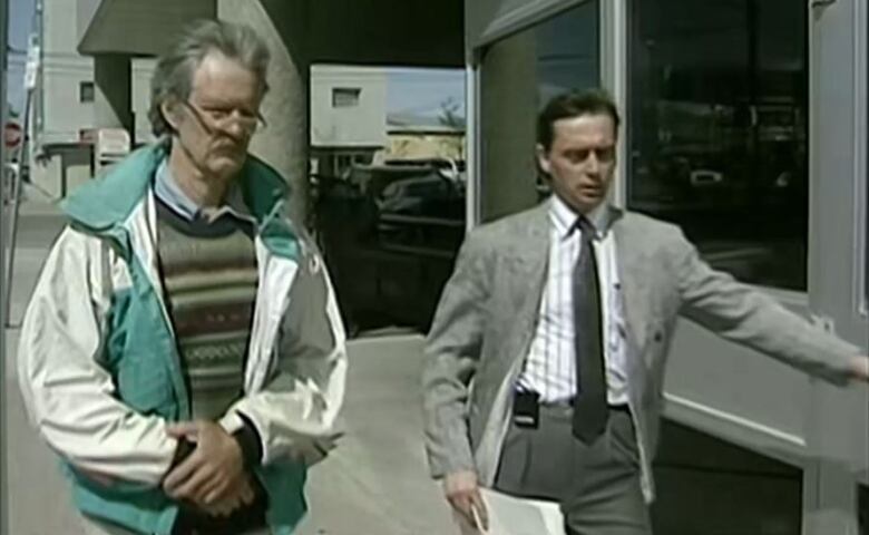 A man grey hair wearing glasses and a striped sweater under a white and green jacket being escorted by his lawyer who is wearing a grey suit