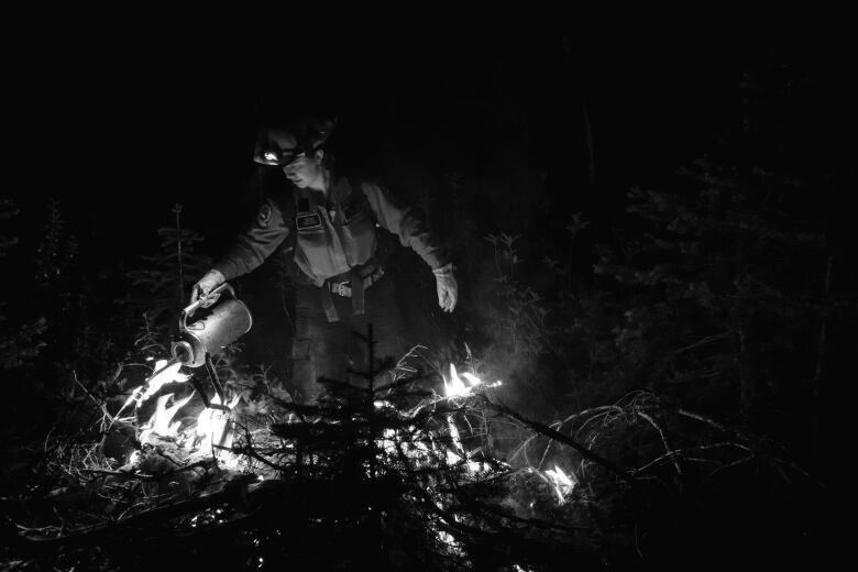 A woman wearing protective gloves holds a canister. Trees and small flames are at her feet.