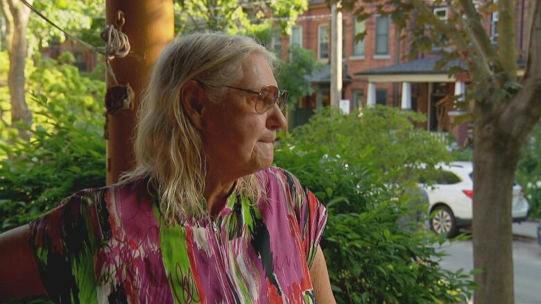 A blond woman wearing sunglass looks past her porch, onto a street. 