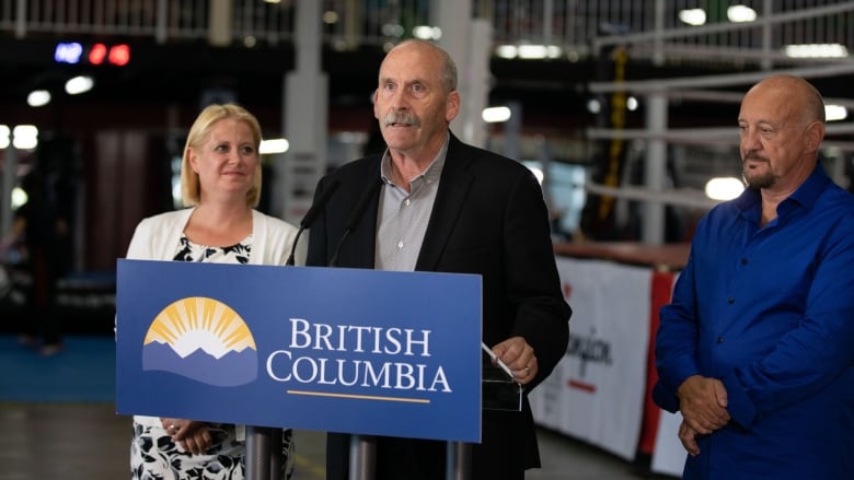 A man speaks at a podium in a boxing gym while a man and woman look on.