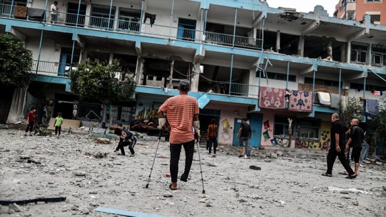 A blue and white concrete building is heavily damaged. People walk in the courtyard strewn with debris