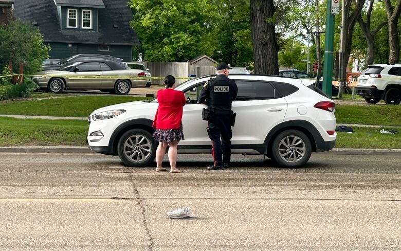 A police officer speaks to a woman standing by a car. A shoe is in the middle of the road.