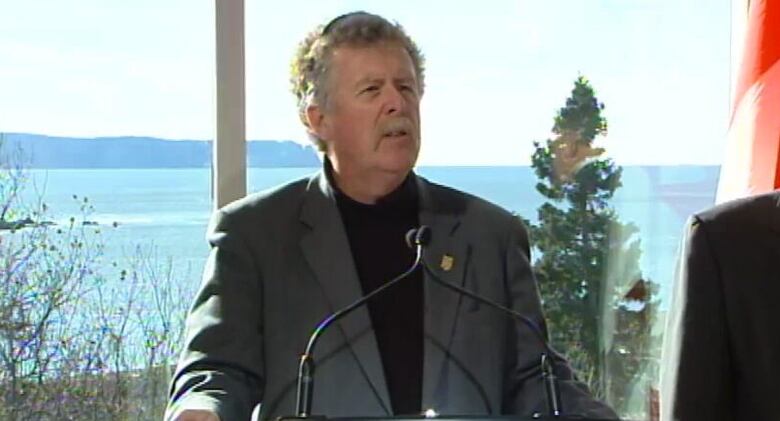 A man wearing a suit speaks into a microphone in front of a window with a view of water behind him. 