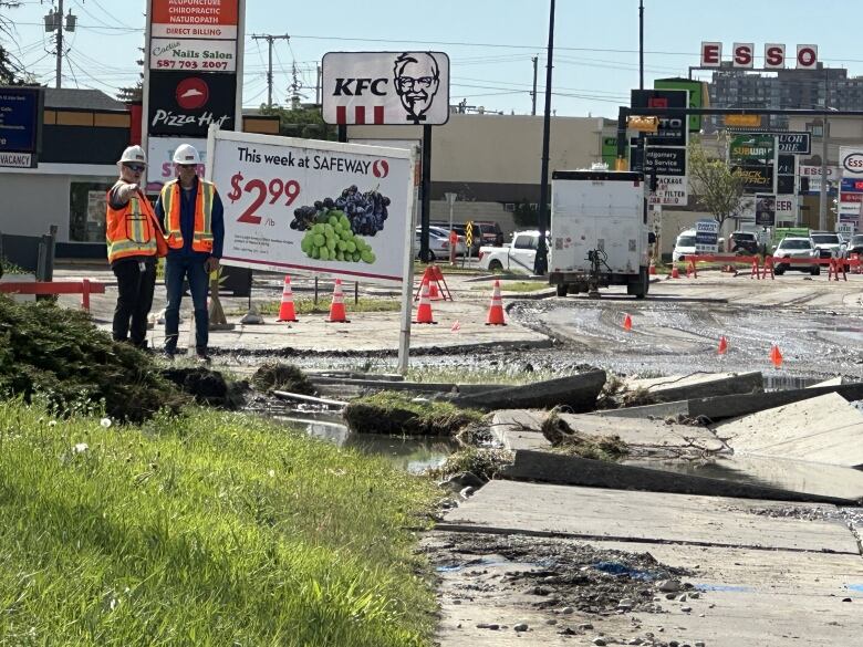 two workers in orange vests point at buckled pavement and a muddy street. 