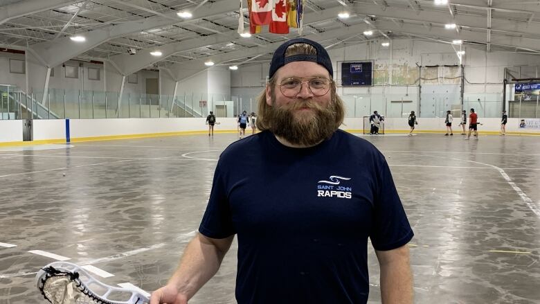 Man with beard holding lacrosse bracket smiling at camera with indoor sports arena in background