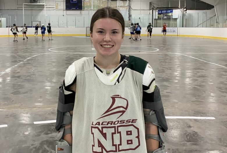 Young woman wearing lacrosse gear smiling at camera with indoor sports arena in background 