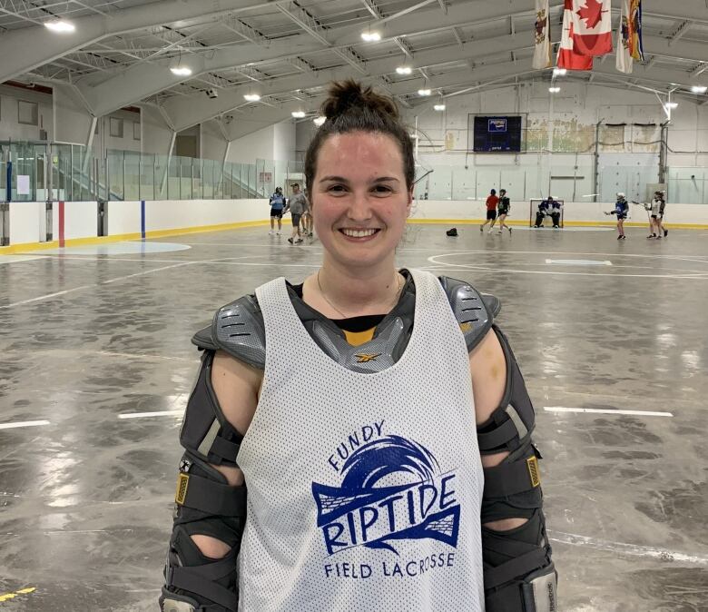 Young woman wearing lacrosse gear smiling at camera with indoor sports arena in background 