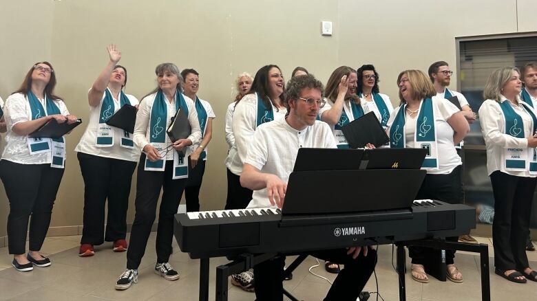 A group of smiling people wearing white tops, black pants and blue scarves gather around a piano, where a man is sitting mid-performance.