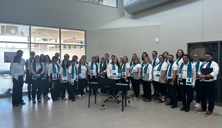 A group of people wearing white tops, blue scarves and black bottoms stand together in front of a piano.