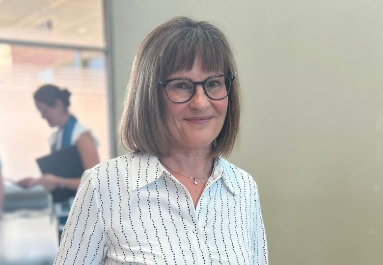 A smiling woman with short brown hair and wearing glasses and a white shirt stands inside an atrium.