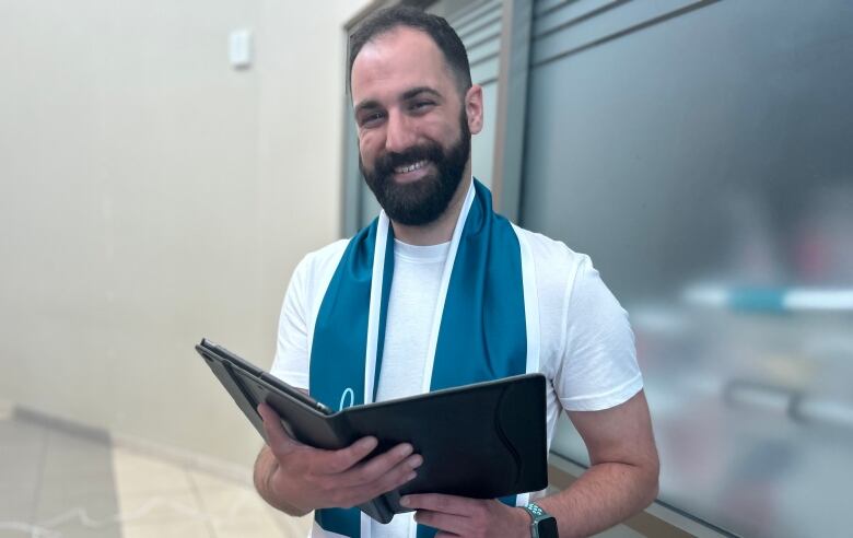 A smiling man with a brown beard and wearing a white t-shirt and blue scarf holds up a black binder.