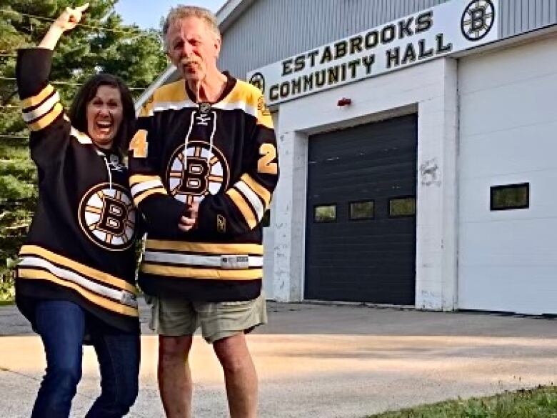 A woman and man wear hockey jerseys and smile outside a community hall.