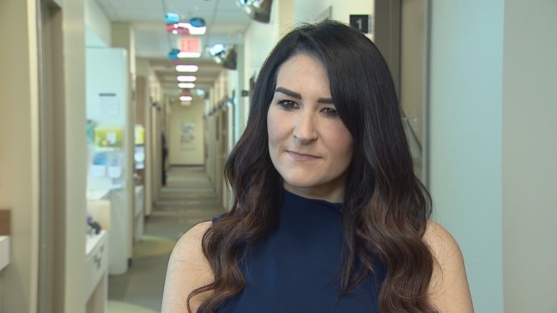 A woman with long dark brown hair is wearing a navy blue dress and standing in the hallway of a medical clinic.