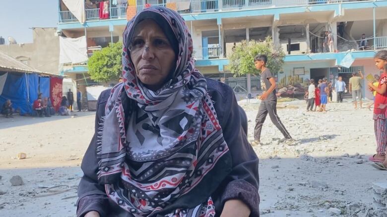 A woman, wearing a red, white, black and grey patterned headscarf, sits outside in front of a damaged building. 