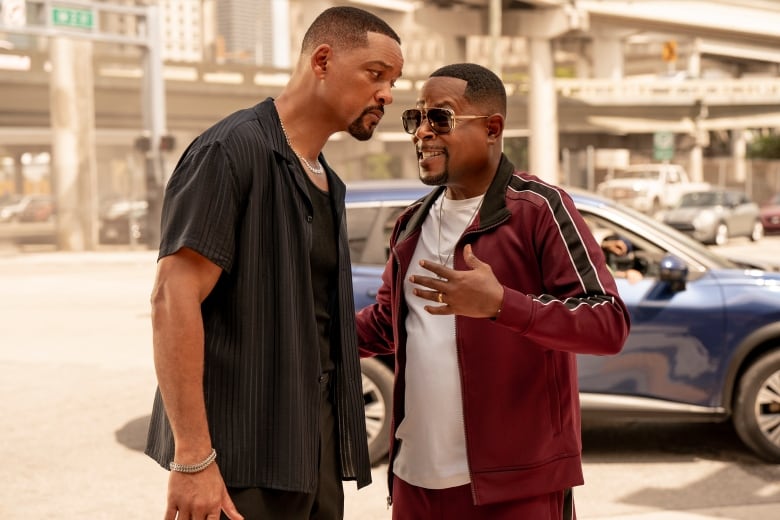 A scowling man, left, listens to a wildly gesticulating man next to him. They are standing outside in the sun.