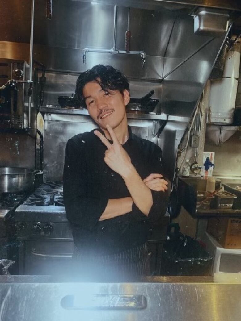 A man holding up a peace sign while standing in a restaurant's kitchen.