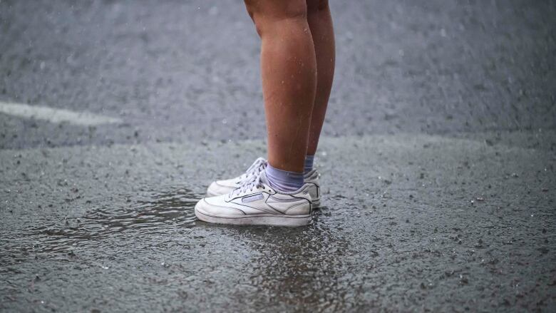 A photo of a person from the knees down. They are wearing sneakers and light purple socks, with no pants we can see. The road is wet around them, with raindrops bouncing off the concrete.