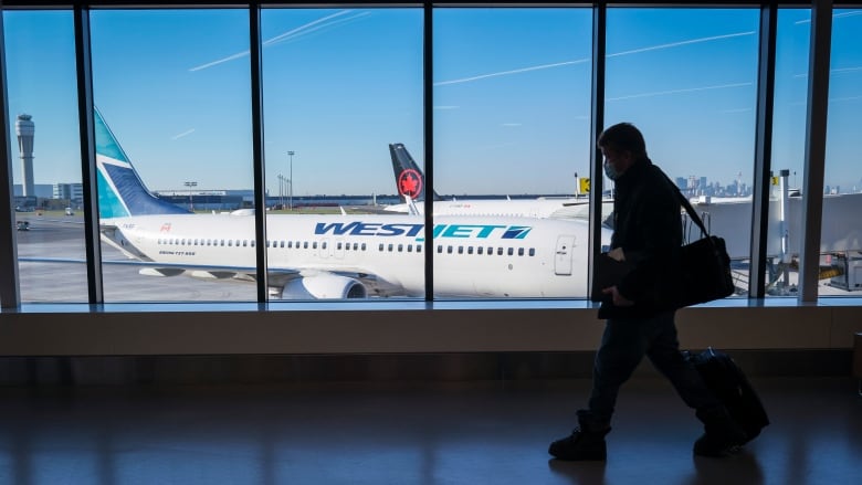 The silhouette of a person walking in an airport terminal in front of  a window with a westjet plane outside.