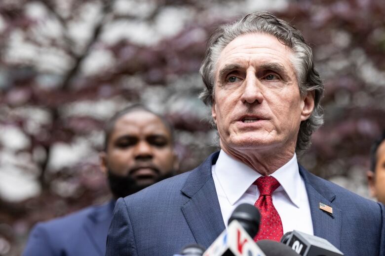 A cleanshaven man with wavy hair in a suit and tie is shown speaking in closeup in an outdoor photo.