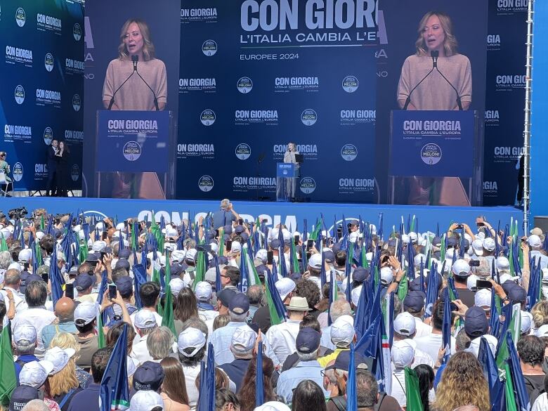 A person speaks to a large crowd as their image is projected twice on a large screen behind them.