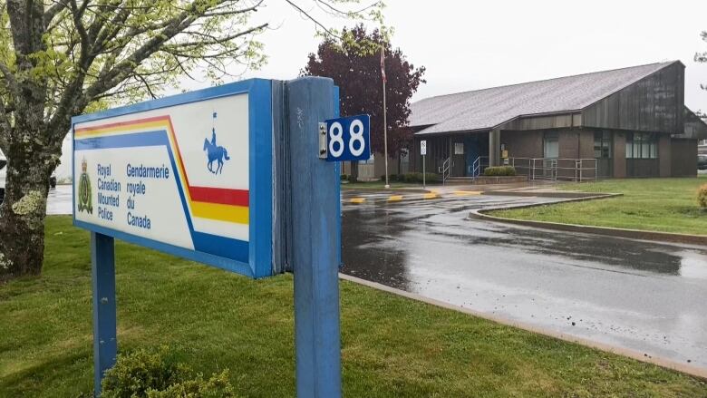 A one-storey brown and grey building is seen behind an RCMP sign with the Mountie crest and red, blue and yellow stripes 