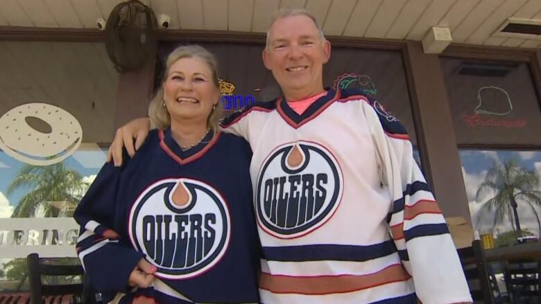 A middle-aged, white couple stand beside each other smiling in the shade. The blonde woman, on the left, is wearing a navy blue Edmonton Oilers jersey. The man with short greying hair, on the right, is wearing a white Oilers jersey.