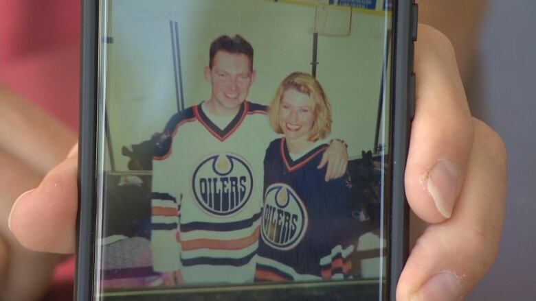 A white man and woman are in the stands of a hockey arena, with the ice behind them. The man, standing on the left, has his arm around the woman. They are both smiling and wearing Edmonton Oilers jerseys.