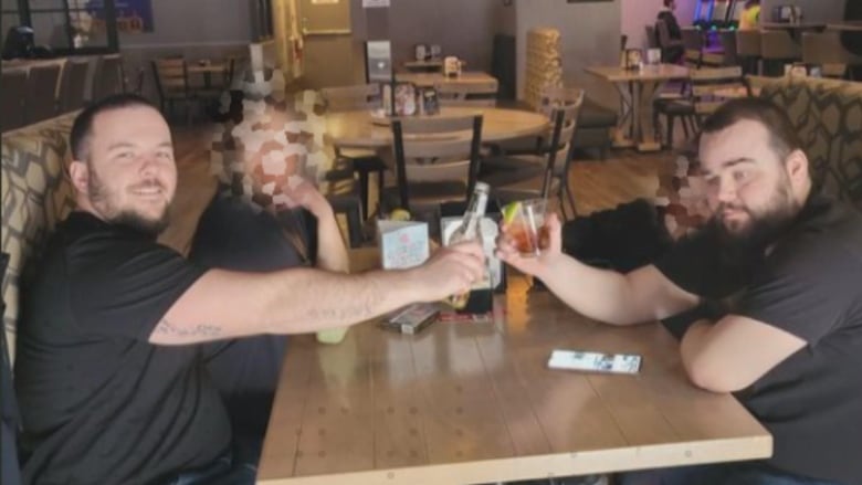 Two men in black t-shirts toast with drinks at a table at a restaurant.