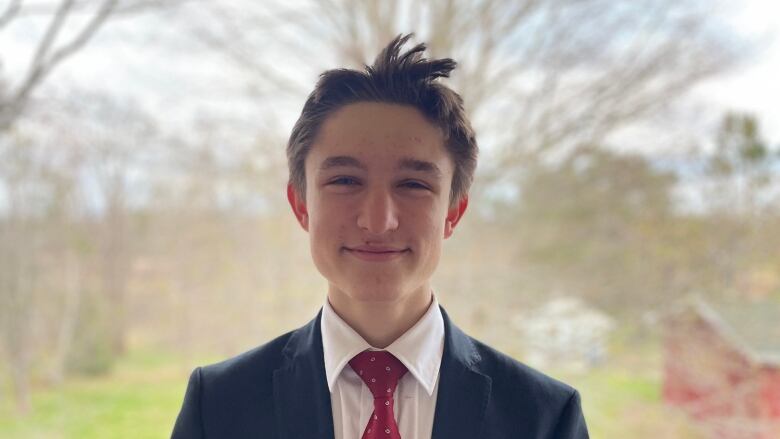 A teenage boy with short hair, wearing a shirt, tie and jacket, stands outside smiling.