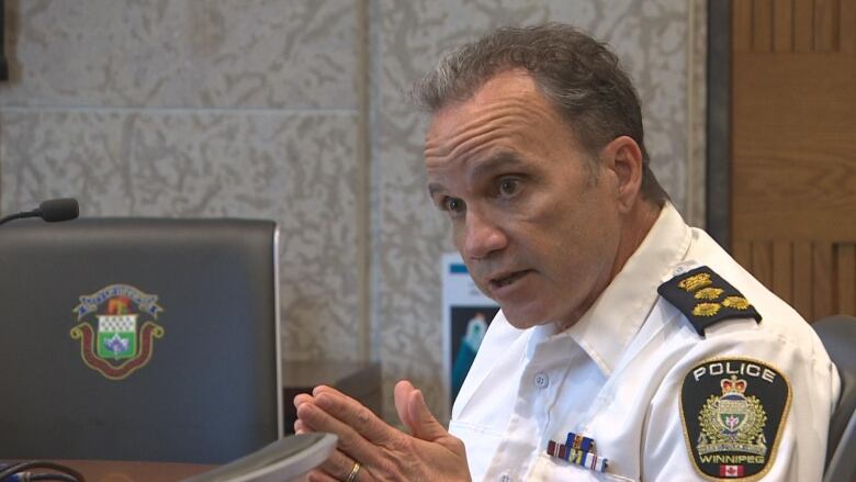 A man wearing a police uniform is sitting in a room next to large wooden table and talking.