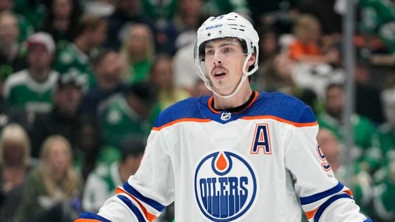 An Edmonton Oilers hockey player is wearing a white uniform on the ice. In the background, fans wearing green Dallas Stars jerseys can be seen in the stands.