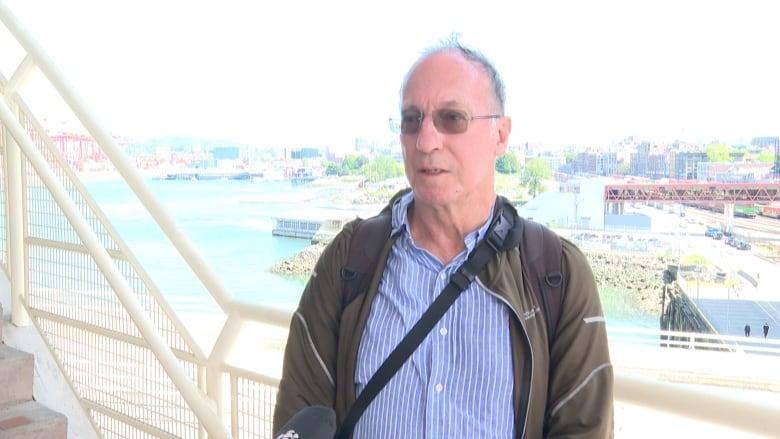 A man donning glasses, a fleece and a jacket interviewed on camera with Vancouver's Coal Harbour in the background. 