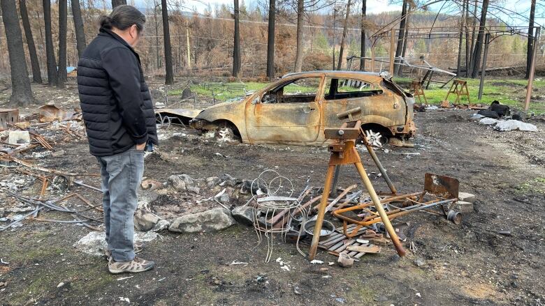 A man wearing a black jacket looks at a burned-out car, metal debris and charred ground.