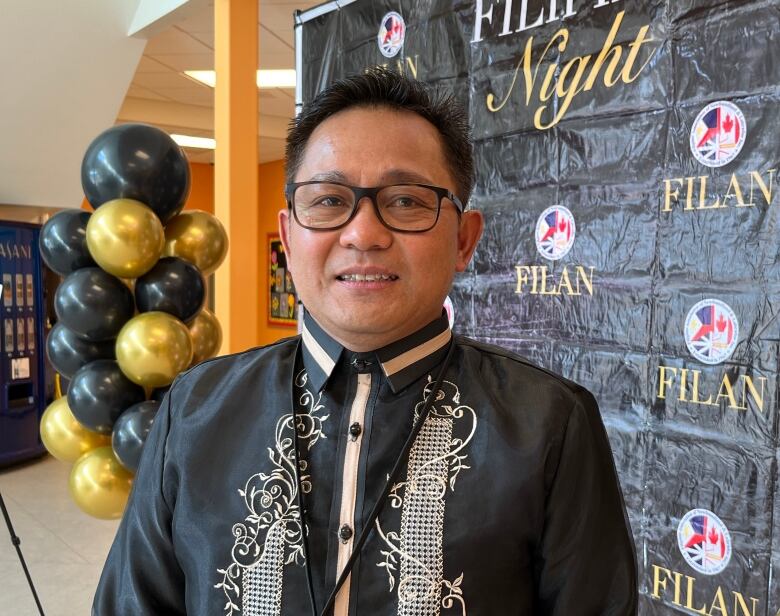 A man smiles. He wears a traditional Filipino shirt, and stands in front of a photo wall that reads FILAN, short for Filipino Association of N.L.