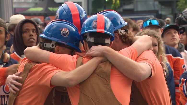 Four men, dressed in blue and orange oil workers uniforms, are huddled together among a crowd outside.