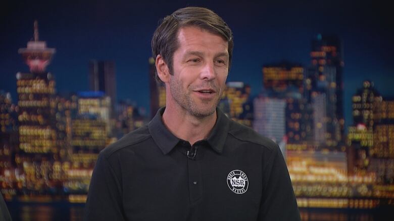 A man with North Shore Rescue logo on his shirt during an interview inside a studio with the Vancouver skyline on the background