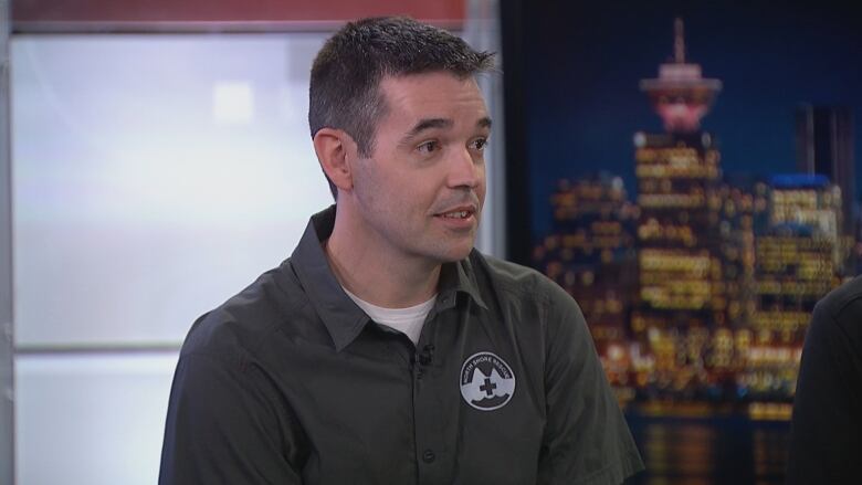 A man with North Shore Rescue logo on his shirt during an interview inside a studio with the Vancouver skyline on the background