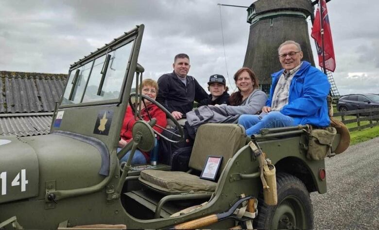 An army jeep in front of a windmill with five people of all ages standing around it. 