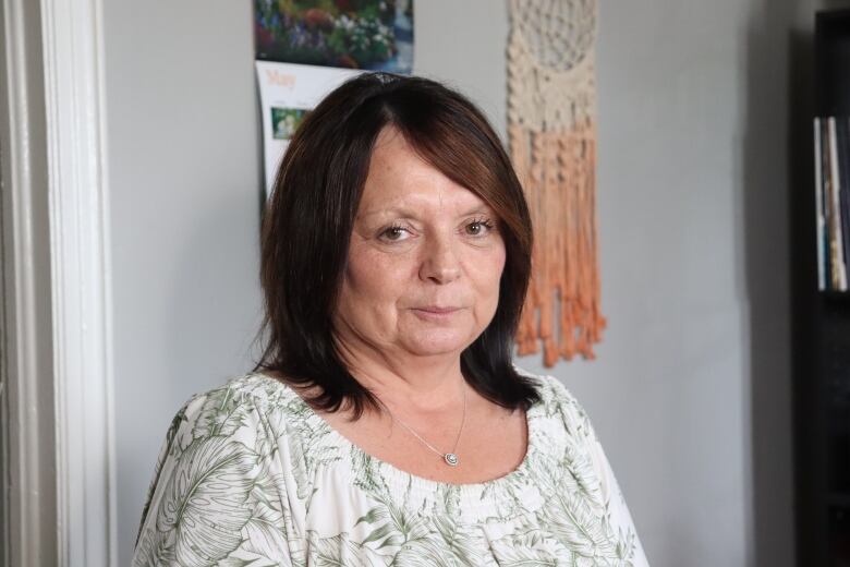 A woman wearing a white patterned shirt standing near a grey wall. 