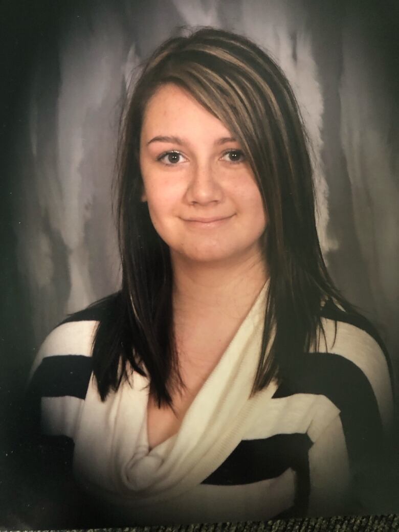 A woman with dark hair and a striped shirt sitting in front of a grey wall. 