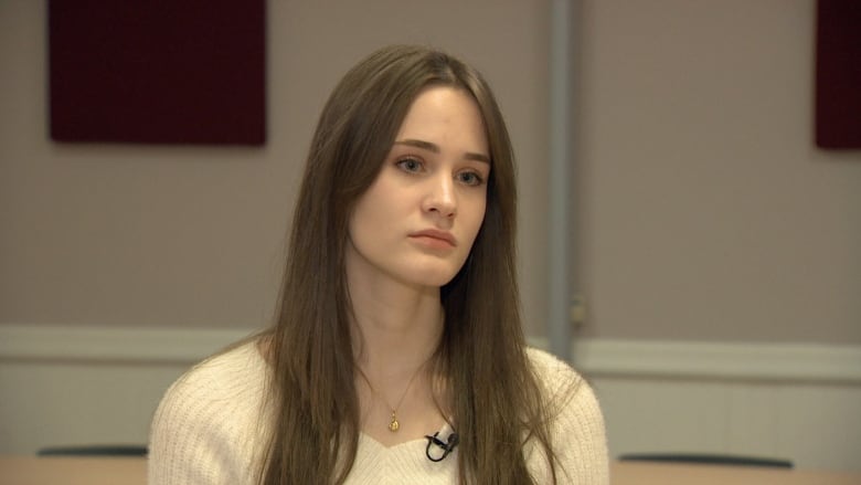 A woman with long brown hair looks somber. She wears a cream sweater and sits in a classroom. 