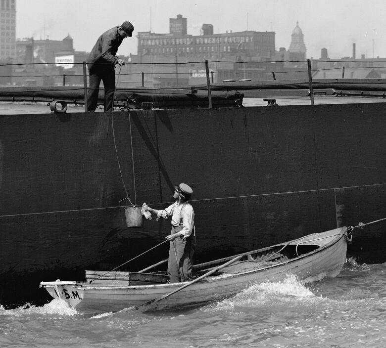 Black and white photo of man standing on rowboat by a large vessel. A rope tied to a bucket is about to be pulled on board.