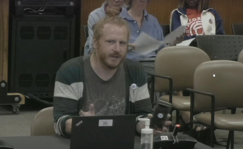 A person with orange-brown hair and a beard speaks in a city council chamber.